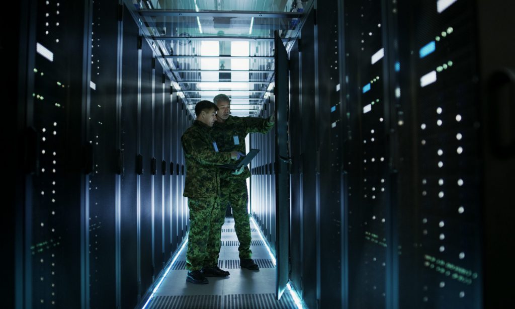 In Data Center Two Military Men Work with Open Server Rack Cabinet. One Holds Military Edition Laptop.